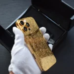 Goldgeprägtes iPhone 16 Pro Max mit kunstvollem Blumenmuster, gehalten in weißen Handschuhen