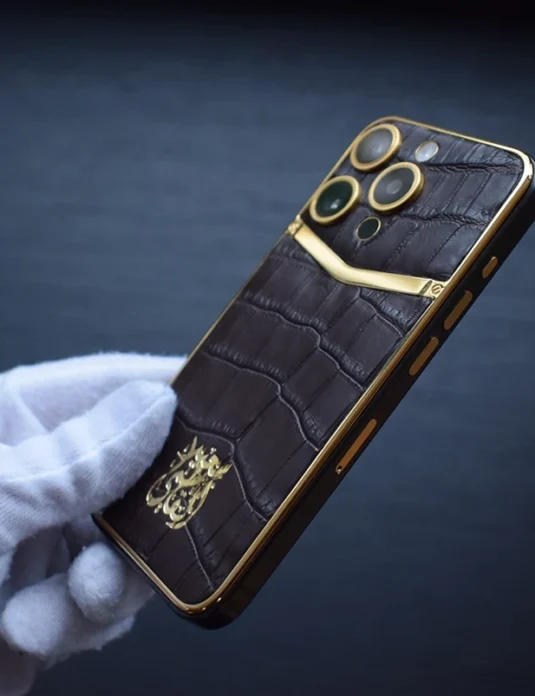 戴着白色手套的手中拿着金色 iPhone 16 Pro，棕色鳄鱼皮机身，纹理清晰可见。