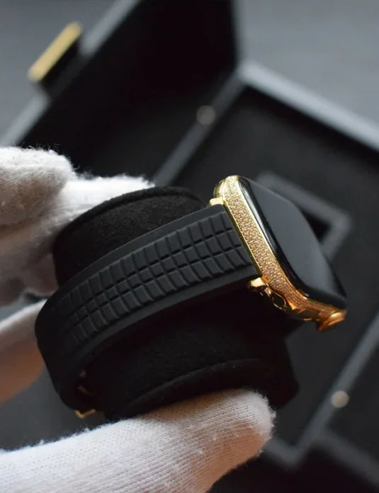 Apple Watch de oro con diamantes, chapado en oro de 24 quilates y bisel engastado con diamantes, con correa de cuero negro, sostenido por manos enguantadas de blanco