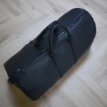 حقيبة سفر جلدية سوداء مزينة بأحرف أولى خضراء منقوشة وتفاصيل سحاب على أرضية خشبية