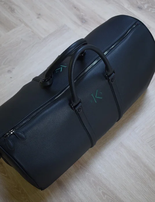 حقيبة سفر جلدية سوداء مزينة بأحرف أولى خضراء منقوشة وتفاصيل سحاب على أرضية خشبية