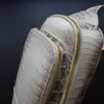 Cream crocodile-textured crossbody bag with gold zippers against a dark background.
