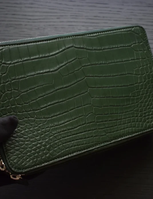 Green crocodile leather pouch with dual zippers and gold hardware on dark wooden surface