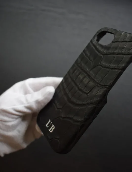 غلاف آيفون تمساح أسود ممسوك بيد مغطاة بالقفاز الأبيض يُظهر نسيج الجلد الطبيعي المتقشر