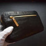 Pochette pour homme crocodile noire tenue dans des mains gantées montrant une finition de cuir texturée et une fermeture éclair dorée
