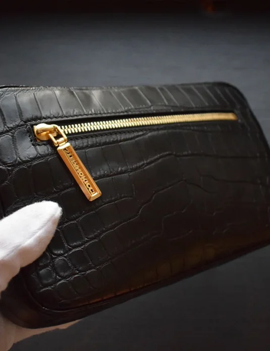Pochette pour homme crocodile noire tenue dans des mains gantées montrant une finition de cuir texturée et une fermeture éclair dorée