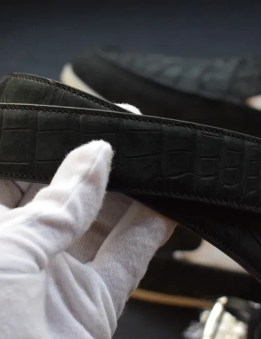 Black crocodile leather belt with distinctive scale pattern being examined with white gloves