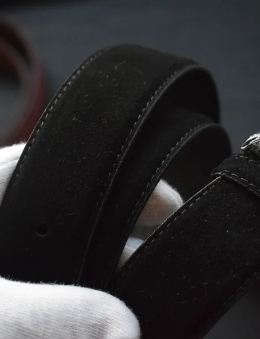 Schwarzer Wildledergürtel mit Premium‑Schnalle, gehalten von behandschuhten Händen, die Handwerkskunst zeigen