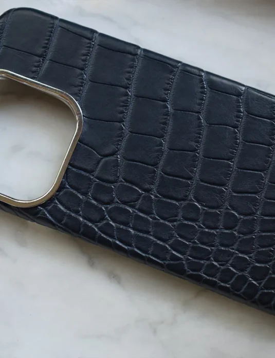 جراب هاتف أزرق بنقشة جلد التمساح مع ملمس جلدي متقشر وحواف ذهبية للكاميرا على سطح رخامي