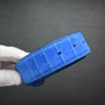 Blue crocodile leather belt with nubuck texture showing natural scale patterns and precision stitching held by gloved hands