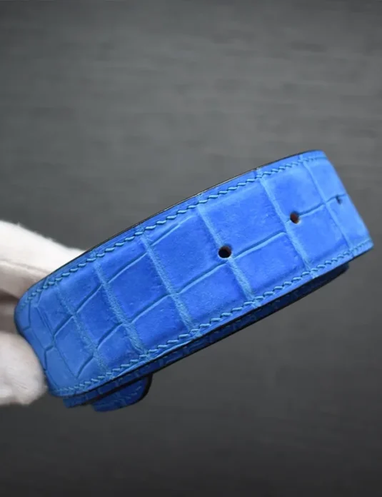Blue crocodile leather belt with nubuck texture showing natural scale patterns and precision stitching held by gloved hands