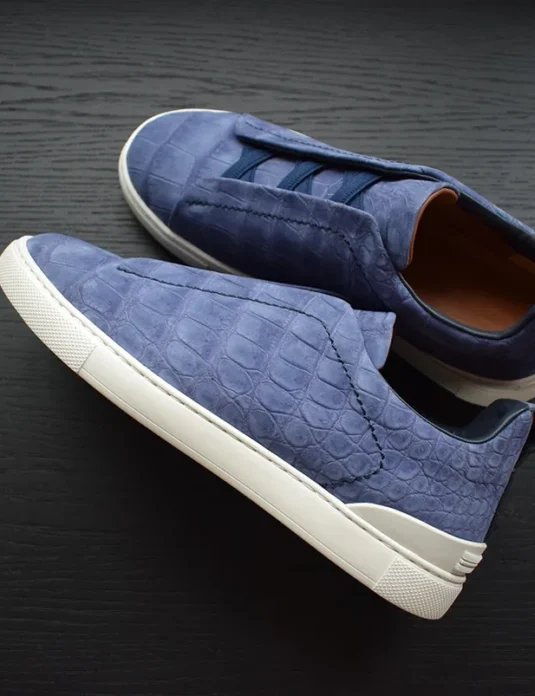 Blue crocodile leather sneakers with triple stitch detailing and white rubber soles on dark wooden surface