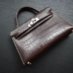 Brown alligator mini kelly handbag with silver hardware and structured design on dark wooden surface