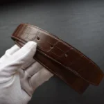 棕色鳄鱼皮皮带，配金色扣环，握于戴白手套的手中，展示纹理鳞片图案