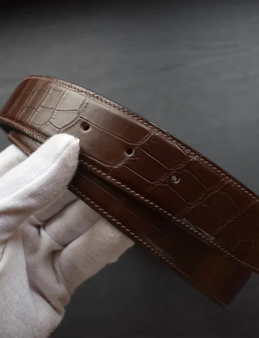 Brown crocodile leather belt with gold buckle held in white gloved hand showing textured scale pattern