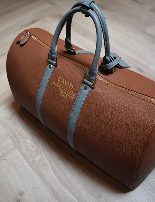 Borsone in pelle marrone con manici grigio-blu su pavimento in legno che mette in risalto i dettagli artigianali