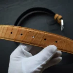 Cognac crocodile leather belt with natural scale texture held by white gloved hands showing craftsmanship details