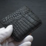 Portatarjetas de cuero de cocodrilo negro sostenido en manos enguantadas blancas que muestran escamas texturizadas y acento dorado