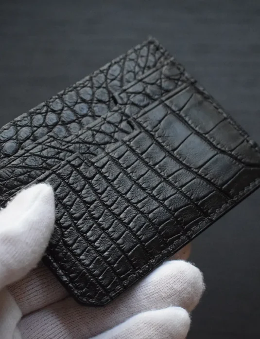 حامل بطاقات من جلد التمساح الأسود يُمسك بأيدي مغطاة باللون الأبيض تُظهر القشور الملمسة واللمسة الذهبية