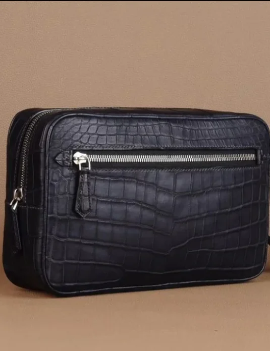 Black crocodile leather clutch with silver hardware and wrist strap on beige background