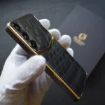 一部金色三星鳄鱼皮手机，黑色鳄鱼皮后盖，24K镀金边框，被一位戴着白手套的人拿在手里。