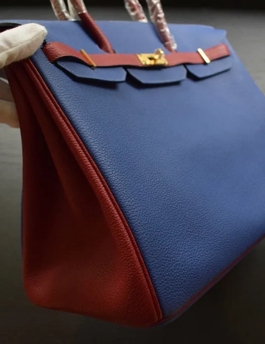 蓝色皮革手袋，Birkin 风格，酒红色内衬和金色五金，由戴手套的手握持