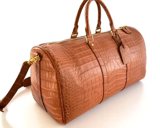 Brown crocodile leather duffle bag with gold hardware and shoulder strap on white background