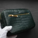 Pochette en cuir de crocodile vert tenue dans des gants blancs montrant une texture écailleuse et des détails de fermeture éclair dorée