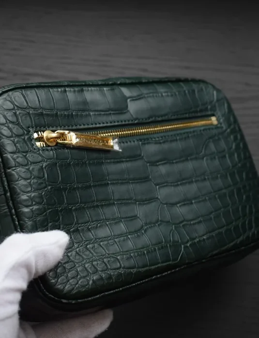 Pochette en cuir de crocodile vert tenue dans des gants blancs montrant une texture écailleuse et des détails de fermeture éclair dorée