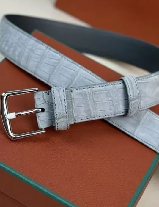 حزام من جلد التمساح الرمادي مع مشبك فضي يعرض حراشف التمساح النوبوك المزخرفة على خلفية برتقالية