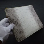 Pochette en cuir de crocodile gris avec des finitions dorées, tenue dans une main gantée de blanc, révélant la texture du crocodile de l'Himalaya