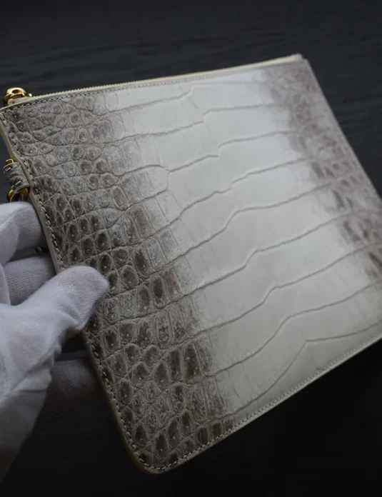 Pochette en cuir de crocodile gris avec des finitions dorées, tenue dans une main gantée de blanc, révélant la texture du crocodile de l'Himalaya