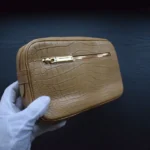 Pochette en crocodile cognac clair tenue dans des mains gantées de blanc montrant le cuir texturé et la quincaillerie dorée