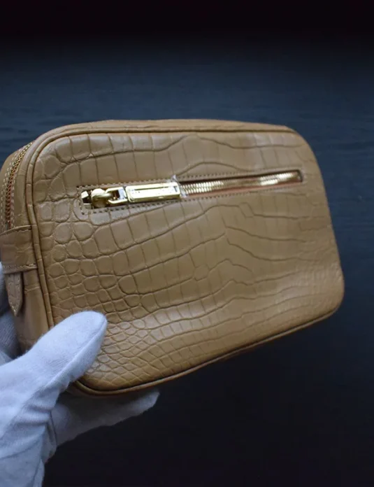 Pochette en crocodile cognac clair tenue dans des mains gantées de blanc montrant le cuir texturé et la quincaillerie dorée