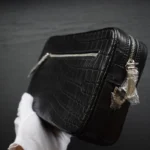 Pochette en cuir de crocodile noir avec fermeture éclair tenue dans des mains gantées de blanc montrant la texture écailleuse