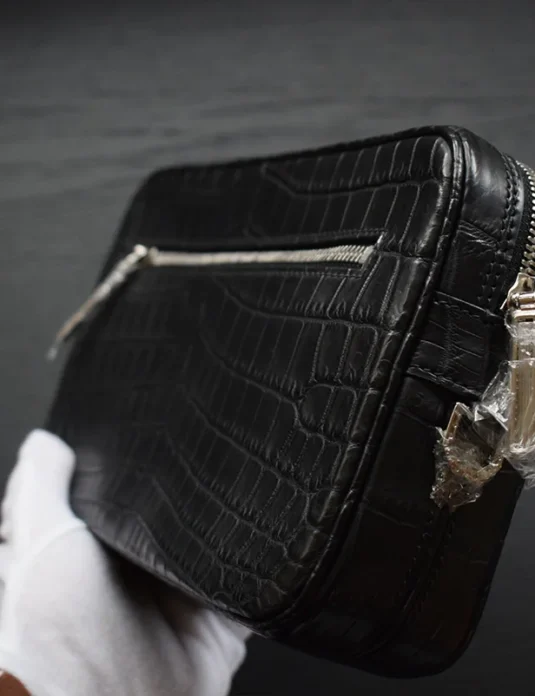 Pochette en cuir de crocodile noir avec fermeture éclair tenue dans des mains gantées de blanc montrant la texture écailleuse