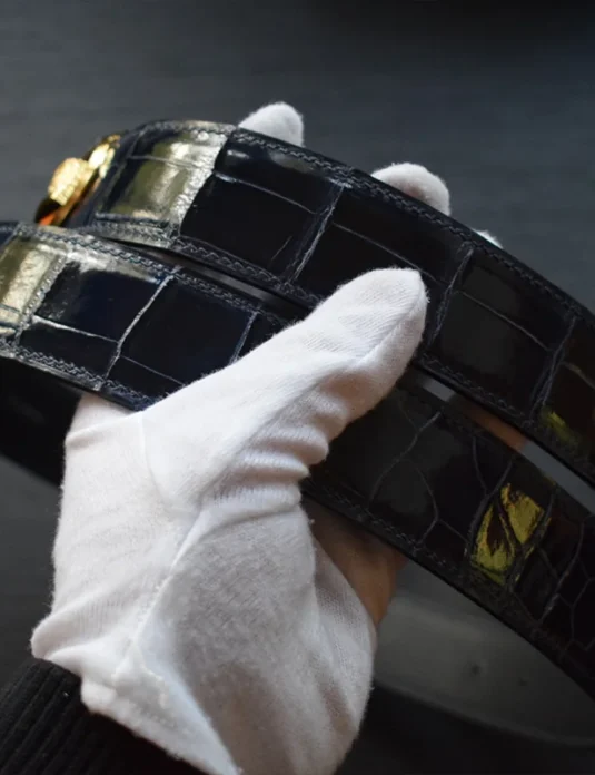 戴着白手套的手中拿着一条蓝色鳄鱼皮腰带，露出独特的鳞片纹理和精湛的工艺。