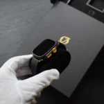 Apple Watch Ultra Gold nero opaco con corona e pulsanti in oro 24 carati, tenuto in mani guantate di bianco