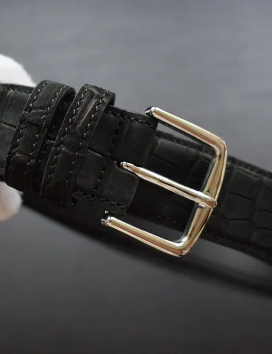 حزام جلدي من تمساح أسود مع مشبك ذهبي يُمسك بأيدي بيضاء مكسوة بالقفاز