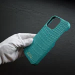 蒂芙尼蓝鳄鱼皮 iPhone 保护壳，白色棉手套握持，展示青绿色皮革纹理