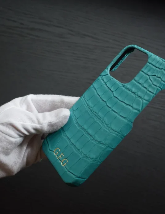 غلاف آيفون من تمساح باللون الأزرق الفيروزي من تيفاني يُحمل بيدين مكسوتين بقطن أبيض يُظهر نسيج الجلد الفيروزي