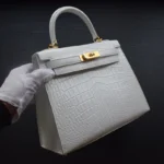 White crocodile kelly handbag held by gloved hands showing structured silhouette and gold hardware