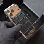 iPhone 17 Pro Max noir façon crocodile avec cadre en or blanc, tenu dans une main gantée, révélant la texture du cuir et la gravure personnalisée